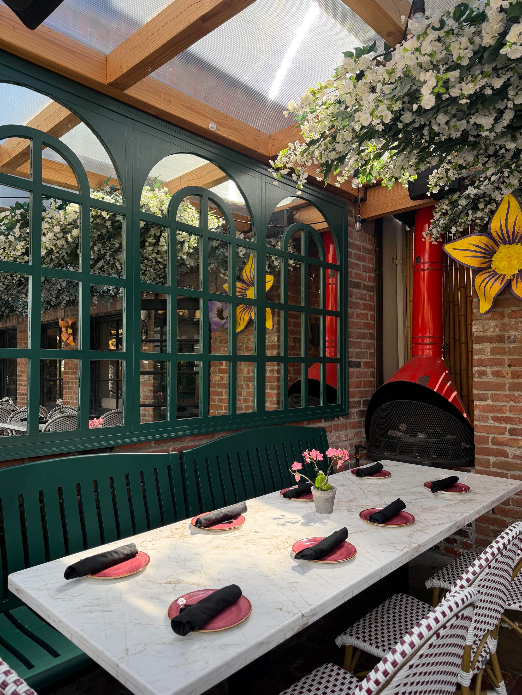 The covered patio: a long marble table set under blossom and a glass roof, green arched windows, brick walls and the red wood-fired oven